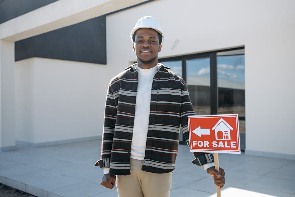Young black male realtor holding a for sale sign in front of a modern house, smiling